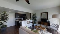 This thoughtfully designed single-floor space showcases a seamless open-concept layout that effortlessly connects the kitchen, dining, and living areas. The kitchen is a standout feature, equipped with sleek dark cabinetry, stainless steel appliances including a range, microwave, and refrigerator, and a light-colored peninsula with bar seating that creates a natural divide between the kitchen and living space. A subway tile backsplash adds a classic touch to the modern aesthetic.

The living area exudes a relaxed yet sophisticated ambiance, anchored by rich dark hardwood flooring that flows throughout the space. A plush, light-toned sofa, rustic wooden accent stools, and a media console with a flat-screen TV contribute to the warm, inviting atmosphere. Natural greenery in the form of a tall indoor palm adds a lively organic element to the room.

The high ceilings, enhanced by a ceiling fan that provides both function and style, lend an airy, spacious feel to the interior. Neutral walls allow the carefully chosen furnishings and natural wood tones to take center stage. Overall, the space offers a modern, comfortable environment with a harmonious blend of contemporary design and natural materials.