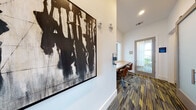 Located at 1040 Jamison Loop in Kissimmee, Florida, this single-floor amenity space is thoughtfully designed to serve as a professional and collaborative environment. The layout includes a corridor-style business center equipped with a long, built-in wooden work surface and ergonomic rolling chairs, providing dedicated spots for individual focus work. Large interior windows along the hallway offer views into an adjacent lounge area, keeping the space feeling open and connected. The patterned carpet throughout — featuring bold striped tones of blue, gold, and grey — adds a contemporary energy underfoot. Private conference rooms branch off from the main corridor, each outfitted with a round white pedestal table, task chairs, and floor-to-ceiling windows dressed with sheer curtains that invite abundant natural light and outdoor views. The rooms are adorned with striking large-scale artwork, including a dramatic black-and-white abstract piece and a bold graphic owl print against textured gold wallcovering, lending each space a distinct creative character. A decorative white laser-cut screen panel adds an architectural accent, visible through interior windows. The overall ambiance balances modern professionalism with artistic warmth, making this a well-appointed space for meetings, focused work, and collaboration.