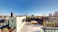 Located in the Westwood neighborhood of Los Angeles, this rooftop terrace offers an expansive open-air experience with sweeping panoramic views of the surrounding cityscape, including recognizable high-rise buildings and the broader LA basin. The deck is finished with large-format porcelain tiles throughout, lending a clean, modern aesthetic to the entire space.

A central lounge area features a deep, curved sectional sofa in dark upholstery paired with a round travertine-topped coffee table, anchored beside a sleek, horizontal linear fireplace set into a stucco accent wall — ideal for evening gatherings. Raised planter beds line the perimeter, filled with drought-tolerant succulents, ornamental grasses, and native plantings that add texture and greenery without obstructing the views.

A dedicated outdoor dining zone accommodates both counter-height bar seating and a round bistro-style table with rope-woven chairs, complemented by an outdoor kitchen or bar station. Elevator access is conveniently available directly on the rooftop level, alongside stairwell entry. Metal railings frame the perimeter, ensuring safety while preserving unobstructed sightlines. Under the clear Southern California sky, this rooftop terrace serves as a well-appointed communal amenity space designed for both relaxation and social entertaining.