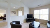 This single-floor space offers a clean and functional layout centered around a well-appointed living area. The room is immediately distinguished by its vaulted ceilings, which lend a sense of openness and airiness to the interior. Neutral, light-toned walls complement the carpeted flooring, creating a warm and inviting atmosphere throughout.

Large windows allow abundant natural light to fill the living area, enhancing the brightness of the space and offering views of the surrounding exterior. A half-wall partition adds an architectural detail that subtly defines different zones within the open layout without sacrificing the sense of spaciousness.

The living area is thoughtfully arranged to accommodate both relaxation and entertainment, with ample room for seating and media setup. Recessed lighting supplements the natural light, ensuring the space remains well-lit at all times of day.

Overall, the space presents a straightforward yet comfortable environment, characterized by its airy ceiling height, generous natural light, and cohesive neutral palette. The practical design and open feel make it a versatile and welcoming living space suitable for a variety of lifestyles.