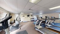 Located at 1401 Eland Downe in Phoenixville, PA, this fitness center offers a comprehensive exercise facility designed to support a full range of workout routines. The space is housed on a single floor and features a drop-panel ceiling with recessed fluorescent lighting that ensures bright, even illumination throughout. Multiple windows along the walls allow natural light to filter in, contributing to an open and energizing atmosphere.

The exercise room is thoughtfully equipped with a diverse selection of cardio machines, including treadmills, elliptical trainers, upright and recumbent stationary bikes, and a stair stepper. For strength training, the facility features a multi-station cable machine and a dedicated free weights area with a full rack of dumbbells in varying sizes. Exercise mats and open floor space provide room for stretching, yoga, and floor-based workouts.

A wall-mounted flat-screen television adds to the workout experience, while a wall-mounted air conditioning unit helps maintain a comfortable indoor climate year-round. The flooring consists of a combination of carpet and rubber exercise mats, providing both comfort and stability across different workout zones. Informational postings and a clock are mounted on the walls, adding functional touches to the environment. Overall, the facility is well-organized and caters to a variety of fitness needs.