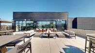 This expansive rooftop terrace is a well-appointed outdoor amenity space defined by its contemporary architectural design and thoughtful layout. The terrace is anchored by a modern building clad in dark horizontal ribbed panels, with floor-to-ceiling glass windows and doors that connect the indoor amenity space to the outdoor deck, allowing natural light to flow freely between the two areas.

The outdoor terrace is finished with large-format concrete pavers that extend across the entire deck, creating a clean, cohesive aesthetic. Multiple seating arrangements are distributed throughout, featuring plush neutral-toned sofas, lounge chairs with warm wood frames, ottomans, and low coffee tables — all designed for comfortable outdoor gathering. Bar-height seating and tables provide additional casual dining options.

A dedicated grilling zone includes multiple stainless steel barbecue stations accompanied by counter-height seating, ideal for outdoor cooking and entertaining. Raised planters filled with ornamental grasses, shrubs, and mature trees add lush greenery and a sense of privacy throughout the space.

Along the perimeter, open railings frame panoramic city views, offering an elevated perspective of the surrounding urban skyline. The overall ambiance is modern, polished, and inviting — designed to serve as a premier outdoor social and relaxation destination.