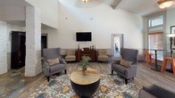 Located in Fort Worth, TX, this single-story community clubhouse at Wildwood Circle North offers a well-appointed gathering space designed for comfort and social interaction. The main lounge area immediately impresses with its soaring vaulted ceilings accented by exposed beams, creating an open and airy atmosphere. The space features rich wood-look flooring that flows throughout, complemented by a bold, multicolored area rug that anchors a central seating arrangement of upholstered wingback chairs and a rustic wood-and-metal coffee table. A wall-mounted flat-screen television and a classic dark wood bookcase add both function and character to the lounge. Distinctive stacked-stone accent columns frame transitions between areas, lending a refined, architectural quality to the interior. Adjacent to the main lounge, a bright café-style room features an array of high windows with wood blinds that flood the space with natural light, along with tall bistro tables and industrial-style bar stools — ideal for casual gatherings. A dedicated dining area with a chandelier provides additional seating, while dark wood cabinetry and curio pieces throughout contribute to the warm, eclectic aesthetic. The overall ambiance is welcoming and thoughtfully designed, balancing comfort with visual interest.