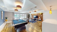 Located at 4041 Radford Ave in Studio City, California, this single-floor amenity space is designed as a modern, multipurpose clubroom that blends entertainment and comfort seamlessly. The open-plan layout is anchored by rich hardwood flooring that flows throughout, unifying the distinct activity zones within the space.

The entertainment area serves as the heart of the room, featuring a full-size pool table positioned beneath stylish drum pendant light fixtures. A bold blue accent wall with framed artwork provides a vibrant backdrop, adding character and depth to the space. Adjacent to the pool table, a well-appointed lounge area offers plush sectional seating, upholstered ottomans, and a patterned blue area rug arranged around a wall-mounted flat-screen television set within a warm wood shelving unit — ideal for gatherings and relaxed viewing.

Large windows invite abundant natural light, offering views of the surrounding streetscape and greenery. Tray ceilings with recessed lighting add architectural detail and a sense of refined height throughout the room.

The kitchen area is thoughtfully equipped with warm wood cabinetry featuring frosted glass inserts, a light-toned tile backsplash in aqua blue, stainless steel appliances, and crisp white countertops. A generous bar-height island with wooden stools provides casual seating, making it well-suited for entertaining. Amber pendant lights add a warm, inviting glow over the prep and dining area.