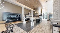 Located at 480 N Orange Ave in the heart of Orlando, FL, this single-floor amenity space offers a seamlessly connected layout designed for both social interaction and everyday convenience. The open-concept kitchen serves as a central showpiece, featuring a large quartz-topped island with integrated sink and bar seating, sleek dark cabinetry, a bold stainless steel range hood, mosaic tile backsplash, open floating shelves, and professional-grade stainless steel appliances — including a built-in coffee station. Teardrop pendant lights cascade above the island, adding an artistic focal point to the culinary space.

The adjacent lounge and dining areas transition effortlessly across mixed-material flooring — a striking combination of wide-plank light wood and dark slate tile. A sculptural textured white wall panel anchors one seating zone, complemented by high-top tables, contemporary bar stools, and a starburst chandelier. The welcoming lounge area features soaring ceilings, floor-to-ceiling sheer drapery, large glazed windows that draw in abundant natural light and views of the streetscape, and a wood-paneled ceiling detail that adds warmth and texture. The overall ambiance is refined, modern, and inviting throughout.
