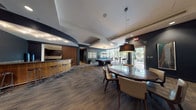 This thoughtfully designed single-floor amenity space offers a diverse range of gathering areas unified by a contemporary aesthetic. The main lounge area is anchored by a large sectional sofa, warm leather armchairs, and a live-edge wood coffee table arranged beneath a series of bold pendant lights. A gallery wall of vibrant blue abstract artwork adds a curated artistic touch, while dark wood-toned flooring runs throughout, grounding the overall design.

Adjacent to the lounge, a billiards table serves as a focal point for recreation, positioned near a wet bar with wood-paneled cabinetry and bar stool seating. A dining area with an oval dark stone table and upholstered chairs provides a more formal gathering spot, positioned close to a sun-drenched nook enclosed by floor-to-ceiling windows that offer lush outdoor views and abundant natural light. High-top tables and modern bucket chairs furnish this bright corner.

The full kitchen is outfitted with dark espresso cabinetry, granite countertops, a stone tile backsplash, and a complete suite of stainless steel appliances, making it well-equipped for catered events or communal use. Throughout, recessed lighting, pendant fixtures, and clean architectural lines contribute to a polished, welcoming atmosphere.