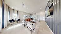 Located at 2935 Ashland Cir in Fort Pierce, Florida, this clubhouse lounge is a single-floor gathering space designed with a clean, contemporary aesthetic. The open-plan living area is defined by crisp white walls, large-format light tile flooring, and recessed lighting that creates a bright, welcoming ambiance throughout.

The room's most striking architectural feature is a floor-to-ceiling dark charcoal slatted accent wall, which serves as a commanding focal point and houses a wall-mounted flat-screen television. Multiple seating arrangements are thoughtfully arranged throughout the space, including plush white accent chairs, a rich cognac leather sofa, and a substantial central table with warm gold-toned metal legs — offering both casual lounging and social gathering options.

A dedicated dining nook near the floor-to-ceiling windows features a round table with dark upholstered chairs, bathed in natural light filtering through elegant navy-striped floor-length curtains. A beverage station with dark cabinetry is conveniently integrated along one wall, adding a functional amenity to the space. Glass double doors open to an outdoor area, reinforcing the connection to the Florida surroundings. The overall atmosphere is polished yet inviting, reflecting a modern Florida lifestyle.