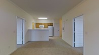 This single-floor residential unit offers a functional and straightforward open-concept layout that connects the living area and kitchen into one cohesive space. The kitchen is positioned centrally, defined by a peninsula counter that provides a natural division between cooking and living areas. It is outfitted with warm-toned wooden cabinetry, a white refrigerator, a built-in microwave, and overhead fluorescent lighting that ensures a well-lit workspace. The countertops are light-colored, complementing the neutral palette throughout the unit. The living area extends generously from the kitchen, finished with wall-to-wall carpet in a light beige tone that adds warmth underfoot. Crown molding runs along the ceiling, lending a subtle architectural detail to the otherwise clean and simple design. The walls are painted in a soft, warm white, contributing to an airy and open ambiance. Two sets of bi-fold closet doors are visible along the adjacent wall, offering ample storage within the living space. A paneled entry door provides access to additional areas of the unit. Overall, the space presents a clean, neutral, and practical environment well-suited for everyday residential living.