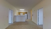 This single-floor residential unit offers a functional and straightforward open-concept layout that connects the living area and kitchen into one cohesive space. The kitchen is positioned centrally, defined by a peninsula counter that provides a natural division between cooking and living areas. It is outfitted with warm-toned wooden cabinetry, a white refrigerator, a built-in microwave, and overhead fluorescent lighting that ensures a well-lit workspace. The countertops are light-colored, complementing the neutral palette throughout the unit. The living area extends generously from the kitchen, finished with wall-to-wall carpet in a light beige tone that adds warmth underfoot. Crown molding runs along the ceiling, lending a subtle architectural detail to the otherwise clean and simple design. The walls are painted in a soft, warm white, contributing to an airy and open ambiance. Two sets of bi-fold closet doors are visible along the adjacent wall, offering ample storage within the living space. A paneled entry door provides access to additional areas of the unit. Overall, the space presents a clean, neutral, and practical environment well-suited for everyday residential living.