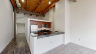 This thoughtfully designed loft-style unit blends industrial character with functional living. The most striking architectural feature is the soaring exposed wood beam ceiling with steel cross-bracing and track lighting overhead, lending the space a warm, warehouse-inspired aesthetic that defines its overall ambiance.

The open-concept layout places the kitchen at the heart of the unit, visible from the main living area below. The kitchen is outfitted with rich cherry wood cabinetry, dark granite-style countertops, stainless steel appliances including a full-size refrigerator, microwave, and electric range, and a deep single-basin sink. Light tile flooring keeps the kitchen bright and easy to maintain. A peninsula counter with bar seating connects the kitchen to the living space, making it ideal for casual dining or entertaining.

The main living area features neutral carpet and flows seamlessly beneath the dramatic ceiling. An arched window brings in natural light and serves as a charming architectural accent visible from the upper level.

An upper-level loft area, accessible by a staircase with a railing, provides a private sleeping or lounging space with carpeted flooring and continued views of the exposed beam ceiling and arched window.

The bathroom is clean and functional, featuring full white tile surround in the tub/shower combination, a vanity with dark countertop, and tile flooring throughout.