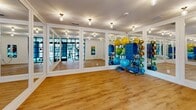 Located at 25 W Seventh Street in Richmond, Virginia, this fitness center is thoughtfully designed to accommodate a full range of workout activities within a single-floor layout. The space is divided into distinct functional zones, each tailored to specific training needs.

The main cardio and strength area features soaring double-height ceilings that create an open, airy atmosphere. Large windows along the exterior wall flood the space with natural light while offering views of the surrounding neighborhood. The walls are accented with bold teal panels, adding a modern, energetic aesthetic. This zone houses multiple treadmills, stationary bikes, elliptical machines, a rowing machine, and a cable machine, alongside a full dumbbell rack and adjustable benches, all set on neutral carpet flooring.

Adjoining the main area is a dedicated studio space finished with warm hardwood flooring and fully mirrored walls framed in crisp white trim. Gold flush-mount light fixtures provide warm, ambient lighting. The studio is stocked with stability balls, medicine balls, resistance bands, and yoga mats, making it ideal for group fitness or functional training.

Steel-framed glass partition doors connect the two zones, maintaining visual openness throughout the facility.