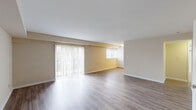 This single-floor unit offers a clean, functional layout designed for comfortable everyday living. The open living and dining area is a highlight of the space, featuring smooth hardwood-style flooring that reflects natural light pouring in through large vertical-blind-covered sliding glass doors that provide views of the surrounding greenery. Neutral warm-toned walls throughout create a bright and welcoming atmosphere.

The kitchen is efficiently designed with rich warm-toned wood cabinetry, light countertops, and a full suite of black appliances including a refrigerator, gas range, built-in microwave, and dishwasher. Light wood-style flooring continues the cohesive aesthetic in this area.

The bedrooms offer plush wall-to-wall carpeting underfoot and feature windows fitted with horizontal blinds, allowing for natural light and privacy. One bedroom includes access to a dedicated closet, providing practical storage.

The bathroom is finished with classic white wall tiles accented by a decorative checkered border in tan and white. It includes a full soaking tub with an overhead shower, a pedestal sink, and a mirrored medicine cabinet with built-in shelving, completing the space with a tidy and functional design.