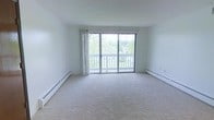 This single-floor space presents a clean, minimalist aesthetic defined by its all-white walls and light neutral carpeting, creating a calm and airy atmosphere. The most prominent feature of the main living area is the large sliding glass door flanked by sheer white curtains, which floods the room with abundant natural light and provides direct access to a private balcony. The balcony overlooks lush greenery and surrounding buildings, offering a pleasant outdoor view. Baseboard heating units run along the perimeter walls, providing an efficient and unobtrusive heating solution. The open layout of the living area allows for flexible furniture arrangements, making the space highly adaptable to various needs. The combination of white walls, soft carpeting, and generous natural light contributes to a spacious and welcoming feel throughout. The simple, understated design of the space ensures that the outdoor views and natural light remain the central focus, enhancing the overall sense of openness and connection to the surrounding environment.