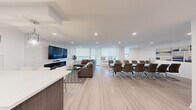 This thoughtfully designed single-floor space offers a seamless open-concept layout that integrates the living, dining, and kitchen areas into one cohesive environment. Light wood-look flooring runs throughout, complementing the crisp white walls and recessed lighting that give the space a bright, contemporary feel.

The living area is anchored by a sleek wall-mounted flat-screen TV above a linear electric fireplace set against a marble-style feature wall. A plush sectional sofa, glass coffee table, and patterned area rug create an inviting lounge setting. A row of roller shade-fitted windows lines the perimeter, allowing abundant natural light while maintaining privacy, with views of outdoor greenery beyond.

Adjacent to the living area, a large conference-style dining table with modern upholstered chairs provides ample seating, making the space well-suited for group gatherings or collaborative use.

The kitchen is outfitted with dark wood cabinetry, white quartz countertops, a subway tile backsplash, and stainless steel appliances including a full-size refrigerator. A generous peninsula with a white quartz waterfall countertop and white bar stools serves as both a prep and casual seating area, illuminated by elegant cylindrical pendant lights.

The bathroom is clean and minimal, featuring white finishes and a shower-tub combination. The overall ambiance is modern, polished, and highly functional.