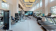Located at 109 W Fortune St in downtown Tampa, Florida, this well-appointed fitness center delivers a comprehensive workout facility designed for serious training. The space is defined by soaring double-height ceilings and expansive floor-to-ceiling windows that flood the interior with natural light while offering views of the surrounding outdoor areas.

The main workout floor is generously proportioned, accommodating an extensive selection of Matrix-branded strength and cardio equipment, including cable machines, multi-functional weight stations, and a full row of treadmills, stationary bikes, and ellipticals. A multi-tiered dumbbell rack holds a complete range of free weights, complemented by dedicated flat and adjustable benches for versatile strength training. Wall-mounted flat-screen televisions are positioned throughout for entertainment during workouts.

A separate enclosed studio or yoga room features glass sliding partition doors with warm wood flooring, providing an intimate and distinct space for stretching, yoga, or personal training sessions. Built-in open shelving within this area conveniently stores stability balls and accessories.

Coffered ceilings with recessed accent lighting and ceiling fans contribute to the polished architectural character of the space, while a bold large-format mural adds a dynamic visual focal point. The light-toned flooring and neutral white walls further enhance the bright, clean ambiance throughout.