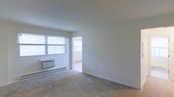 This single-floor residential unit offers a clean, functional layout designed for comfortable everyday living. The main living area is characterized by neutral white walls and soft, wall-to-wall carpeting that creates a warm and inviting atmosphere. Large windows along one wall allow abundant natural light to fill the space, contributing to an open and airy feel. A wall-mounted air conditioning unit and baseboard heating ensure year-round climate comfort. The room opens through a doorway into an adjacent area, suggesting a connected and practical floor plan that allows for easy flow between spaces. The overall aesthetic is minimal and move-in ready, with a bright, clean palette that provides a versatile backdrop for a variety of interior styles. The unit's straightforward design emphasizes functionality, making it well-suited for those seeking a practical and low-maintenance living environment.