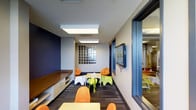 This single-floor facility is thoughtfully designed with children in mind, combining functionality with a fun and welcoming atmosphere. The main activity room is immediately eye-catching, featuring bold navy blue accent walls that contrast with the bright, neutral ceiling. Flush skylight panels and overhead fluorescent lighting ensure the space is consistently well-lit throughout the day. A built-in wooden bench with white lower cabinetry runs along one wall, providing ample storage and seating. The room is furnished with playful, colorful chairs and tables in vibrant shades of orange, green, and white, scaled specifically for young users. Whimsical animal-shaped stools and bean bag chairs add a lighthearted touch to the seating arrangements. A wall-mounted flat-screen display serves as a media focal point, while a glass block window panel introduces diffused natural light and architectural interest. Patterned carpet tiles in muted tones ground the space and provide a durable, comfortable surface for activity. A large sliding glass door connects this room to an adjacent area, which appears to contain additional equipment or activity stations, supporting a seamless flow between spaces. Overall, the facility presents a safe, stimulating, and well-organized environment designed to engage and inspire children.