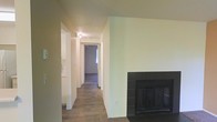 This single-story space is designed with a straightforward, functional layout that connects its rooms through a central hallway. The living area is anchored by a notable fireplace framed in dark tile, serving as a focal point within the space. Neutral, light-toned walls run throughout, contributing to a clean and understated aesthetic.

The hallway extends from the main living area toward the bedrooms, with overhead lighting ensuring the corridor remains well-lit. Wood-style flooring runs through the main areas, offering durability and a cohesive visual flow, while carpeting appears in at least one of the rooms visible at the end of the hall. The transition between flooring materials subtly delineates the living and sleeping areas.

The walls are finished in soft, muted tones that complement the darker flooring, creating a balanced contrast. White trim and baseboards accent the perimeter of each room, adding a degree of architectural detail. A doorway at the end of the hallway allows natural light to filter into the corridor, brightening the interior. Overall, the space presents a practical and neutral environment well-suited for comfortable everyday living.