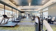 Located in Colorado Springs, CO, this well-appointed fitness center offers a generously sized single-floor layout designed to support a full range of workout routines. The space is divided into two distinct functional zones: a cardio area and a strength training area, each thoughtfully equipped and designed for an optimal exercise experience.

The cardio zone features an impressive lineup of treadmills, ellipticals, and stair climbers arranged along floor-to-ceiling glass walls that flood the space with natural light and offer open views of the surrounding exterior. Large-format mirrors amplify the sense of space, while wall-mounted televisions provide entertainment during workouts. Ceiling fans suspended from the coffered tray ceiling ensure comfortable air circulation throughout.

The strength training area is equally well-equipped, housing cable machines, a squat rack, weight benches, free weights, and a multi-functional cable crossover station. A bold geometric mural in teal, white, and yellow anchors this zone, adding an energetic visual focal point. High clerestory windows provide additional natural light in this area.

Throughout the facility, the flooring consists of multi-toned modular carpet tiles in green, blue, and grey, complementing the cool grey and teal wall palette. Recessed and linear LED lighting, combined with the coffered ceiling design, lend the space a polished, modern ambiance well-suited to an active community fitness facility.