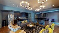 This single-story clubhouse is designed with a contemporary aesthetic, combining bold color accents, polished concrete flooring, and eye-catching circular pendant light fixtures that serve as focal points throughout the space. The main lounge area is generously proportioned, furnished with plush sofas, accent chairs in teal and chartreuse, and patterned area rugs that define distinct seating zones. A full kitchenette runs along one wall, outfitted with dark espresso cabinetry, granite-style countertops, stainless steel appliances including a French door refrigerator, a microwave, and a coffee station, providing convenient amenities for residents and guests. An adjacent secondary lounge benefits from large floor-to-ceiling windows that bring in ample natural light and offer views of the surrounding streetscape, complemented by a vibrant purple accent wall and curated artwork. A separate bar-height gathering area features high-top tables and chairs, a wall-mounted television, a built-in counter, and a glass roll-up garage door that opens directly to an outdoor patio, seamlessly blending indoor and outdoor entertaining. A fitness center is also accessible from within the clubhouse. Throughout, the design maintains a cohesive, urban-modern character.