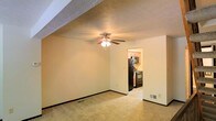 This residential space spans three floors, offering a well-organized layout designed for comfortable everyday living. The main living area features wall-to-wall carpeting that adds warmth underfoot, complemented by a ceiling fan with an integrated light fixture that provides both air circulation and ambient lighting. A textured ceiling adds a classic touch to the room, while neutral-toned walls create a clean, versatile backdrop throughout.

An open doorway connects the living area directly to the kitchen, providing a practical and seamless flow between the two spaces. The kitchen is outfitted with black appliances, including a full-size refrigerator and a gas range, set against warm wood-toned cabinetry that extends to the upper wall. The kitchen flooring transitions to a lighter tile, distinguishing it clearly from the carpeted living space.

Floor vents are integrated into the baseboards, indicating a central heating and cooling system throughout the home. The neutral color palette and straightforward layout make this space highly adaptable to a variety of interior styles. Spanning three floors, the property offers ample room for living, storage, and additional functional spaces, making it a practical and versatile residential option.