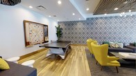 This community room is a thoughtfully designed social and recreational space that seamlessly blends playful functionality with contemporary style. The room features warm hardwood-style flooring that anchors the space and complements the clean white walls and recessed ceiling lighting, which together create a bright, welcoming atmosphere.

A centerpiece ping-pong table by Killerspin occupies the main area of the room, providing an engaging activity for residents and guests. Along one wall, a large mounted Scrabble board offers a classic gaming option alongside a chalkboard, adding a casual and interactive element to the space.

A striking geometric blue-and-white patterned accent wall serves as a bold visual focal point, adding depth and personality to the room's design. Adjacent to this feature wall, a pair of vibrant yellow armchairs provide comfortable seating, offering a relaxed lounge area ideal for socializing or spectating. A tall indoor plant introduces a natural, refreshing touch to the interior.

The combination of recessed lighting and the open layout ensures the space feels both energetic and inviting, making it well-suited for community events, casual gatherings, and recreational activities throughout the day.
