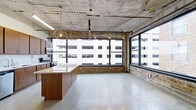 This single-floor space showcases a striking industrial aesthetic, blending raw architectural elements with modern finishes to create a distinctive and functional environment. The exposed concrete ceiling with visible structural details and utility conduits establishes an authentic urban character, while polished concrete floors extend throughout, providing a clean, cohesive foundation underfoot.

A bank of large, black-framed windows along one wall allows abundant natural light to pour into the space, offering open views of the surrounding cityscape. Beneath the windows, original brick masonry adds warmth and texture, creating a compelling contrast against the raw concrete above and below.

The kitchen area is thoughtfully designed, featuring rich dark wood cabinetry, stainless steel appliances including a range and microwave, and a generously sized island topped with a light quartz surface. Geometric pendant lights suspended above the island introduce a contemporary design element that complements the industrial backdrop.

The open-plan layout beyond the kitchen offers considerable flexibility, with the polished floors and expansive footprint accommodating a variety of uses. Track lighting integrated into the ceiling provides supplemental illumination throughout. Overall, the space delivers a well-balanced combination of rugged industrial character and refined modern upgrades.