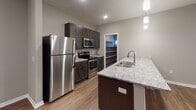 This single-floor residence is designed with a contemporary aesthetic, combining functional layouts with modern finishes throughout. The open-concept living and kitchen area serves as the heart of the unit, where warm wood-look plank flooring extends across a generous, light-filled space. A large kitchen island with a granite countertop and undermount sink anchors the area, complemented by dark espresso cabinetry, stainless steel appliances — including a refrigerator, electric range, and microwave — and pendant lighting overhead. Recessed lighting further brightens the space. A walk-in pantry with built-in shelving adds practical storage to the kitchen area.

The living area opens to an exterior door with an adjacent window, allowing natural light to filter in. The bedrooms are carpeted and well-sized, each featuring ceiling fans and roller shade window treatments. One bedroom benefits from dual windows, providing additional natural light.

Two bathrooms serve the unit. One features a tub-shower combination with chrome fixtures and a dark-cabineted vanity, while the other includes a similarly styled vanity with a white countertop. Wood-look flooring carries through the bathrooms and hallways, maintaining a cohesive design. Neutral gray wall tones throughout create a clean, move-in-ready atmosphere.