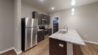 This single-floor residence is designed with a contemporary aesthetic, combining functional layouts with modern finishes throughout. The open-concept living and kitchen area serves as the heart of the unit, where warm wood-look plank flooring extends across a generous, light-filled space. A large kitchen island with a granite countertop and undermount sink anchors the area, complemented by dark espresso cabinetry, stainless steel appliances — including a refrigerator, electric range, and microwave — and pendant lighting overhead. Recessed lighting further brightens the space. A walk-in pantry with built-in shelving adds practical storage to the kitchen area.

The living area opens to an exterior door with an adjacent window, allowing natural light to filter in. The bedrooms are carpeted and well-sized, each featuring ceiling fans and roller shade window treatments. One bedroom benefits from dual windows, providing additional natural light.

Two bathrooms serve the unit. One features a tub-shower combination with chrome fixtures and a dark-cabineted vanity, while the other includes a similarly styled vanity with a white countertop. Wood-look flooring carries through the bathrooms and hallways, maintaining a cohesive design. Neutral gray wall tones throughout create a clean, move-in-ready atmosphere.