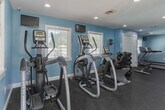 Located at 200 Red Cedar Ct in Chesapeake, Virginia, this fitness center is a well-equipped, single-floor facility designed to support a comprehensive workout routine. The space is finished with calming light blue walls, white trim, and durable rubber flooring, creating a clean and energizing environment throughout.

The cardio area features multiple Matrix treadmills and elliptical cross-trainers, each equipped with individual display monitors mounted on the wall above for entertainment during workouts. Natural light filters in through several white-trimmed windows fitted with blinds, lending the space a bright and welcoming atmosphere.

The strength training section includes a multi-functional cable machine with a variety of attachments, a weight bench, and a full rack of dumbbells ranging across multiple weight increments. A stability ball rounds out the free-weight area, accommodating a range of exercise preferences.

Additional amenities include a wall-mounted water fountain for convenient hydration, hand sanitizer dispensers and posted sanitization notices promoting cleanliness, and a small utility table stocked with cleaning supplies. Recessed lighting supplements the natural light, ensuring the space is well-lit at all times. A wall-mounted television provides additional entertainment options. The facility is serviced by a central HVAC system and is accessible via a standard entry door.