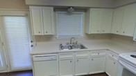 This two-floor space offers a practical and well-organized layout designed for comfortable everyday living. The kitchen is a standout area, featuring classic white raised-panel cabinetry that extends both above and below the counter, providing generous storage throughout. Light-colored countertops run the length of the workspace, offering ample room for meal preparation. A stainless steel double-basin sink is centrally positioned beneath a window fitted with horizontal blinds, allowing natural light to filter into the space while maintaining privacy. A white dishwasher is seamlessly integrated into the lower cabinetry, contributing to the kitchen's cohesive and tidy aesthetic. Under-cabinet lighting above the window adds a functional accent to the space. The overall color palette of white and neutral tones creates a clean, bright atmosphere throughout the kitchen. Spanning two floors, the property provides a sense of spaciousness and separation between living areas, making it well-suited for a variety of needs. The straightforward design and neutral finishes throughout offer a versatile foundation that can be easily personalized to suit individual tastes and preferences.