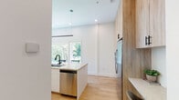 Located at 313 SE Second St in Bentonville, Arkansas, this single-story space showcases a clean, contemporary design characterized by white walls, light hardwood flooring, and warm wood-toned cabinetry throughout. The open-concept kitchen serves as a central highlight, featuring floor-to-ceiling natural wood cabinetry, stainless steel appliances, a subway tile backsplash, and a generously sized island with a stone countertop, undermount sink with a matte black faucet, and seating for multiple guests. A linear pendant light anchors the space, while large windows invite abundant natural light and outdoor views. A dedicated built-in desk area off the kitchen provides a functional workspace. The living area is bright and welcoming, with large glass doors opening directly to an outdoor space overlooking lush greenery. The bedroom continues the refined aesthetic with tall ceilings, hardwood floors, and direct access to the outdoor area through floor-to-ceiling glass doors. The bathroom features a floating wood vanity with matte black fixtures, a round mirror, and a glass-enclosed walk-in shower with large-format tile. An in-unit stacked washer and dryer complete this well-appointed space.