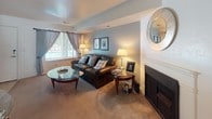 This two-story residence offers a thoughtfully arranged layout across multiple rooms, each finished with practical details and a warm, livable ambiance. The living room provides a comfortable gathering space with soft carpeted flooring, a subtle tray ceiling detail, and a large window dressed with patterned curtains that allow generous natural light. A fireplace surround adds architectural character to the room.

The kitchen is equipped with warm oak cabinetry, light-toned countertops, and white appliances including a range, over-the-range microwave, and dishwasher. A ceiling fan overhead and a peninsula counter add functionality, while blue-toned accent walls contribute a fresh visual contrast.

The bedrooms are carpeted and well-proportioned. One features soft sage-green walls and a window with outdoor views, while another offers a more neutral palette with vertical blinds and ample floor space. A third bedroom includes a closet and window with patterned drapes. Each bedroom conveys a calm, restful character.

The bathroom features tile flooring, an extended vanity countertop with a wide mirror, and chrome fixtures, providing a clean and practical finish throughout.
