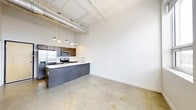 This single-floor loft embraces an industrial-modern aesthetic, blending raw architectural elements with contemporary finishes to create a striking and functional living space. Polished concrete floors stretch across the open-plan layout, reflecting light and reinforcing the loft's urban character. Soaring ceilings draw the eye upward, where exposed ductwork and structural details are left intentionally visible, adding an authentic industrial charm. The kitchen is a central feature of the space, designed with dark wood cabinetry that contrasts elegantly against a clean white countertop. A generous kitchen island with a built-in sink serves as both a practical workspace and a natural social gathering point. Stainless steel appliances — including a refrigerator, microwave, and range — complete the kitchen's modern, functional appeal. A row of pendant lights hang above the island, casting warm, focused illumination and adding a refined touch to the otherwise raw aesthetic. The open living area beyond the kitchen offers a versatile, expansive canvas, with crisp white walls providing a neutral backdrop ideal for a variety of interior styles. The seamless flow between the kitchen and living zones enhances the sense of openness and makes the space well-suited for both everyday living and entertaining.