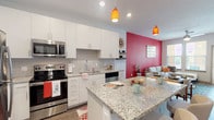 This single-floor residence showcases a thoughtfully designed open-concept layout that seamlessly connects the kitchen, dining, and living areas. The kitchen is a standout feature, equipped with stainless steel appliances including a refrigerator, range, and built-in microwave. Crisp white cabinetry pairs with granite countertops and a classic subway tile backsplash, while a central island provides additional prep and seating space. Warm pendant lighting in amber tones adds a welcoming glow throughout.

The living area is anchored by a bold red accent wall and complemented by wood-look flooring, a ceiling fan, and windows that bring in natural light with outdoor views. A separate entryway area features its own door with sidelight windows and wood-look flooring, creating a defined transition space.

The bedroom offers a comfortable, carpeted retreat with large windows, ample natural light, and room for a full furniture arrangement. A second bedroom or flexible space is also present within the layout.

The bathroom is well-proportioned, featuring a granite-topped double vanity with white cabinetry, wood-look flooring, and a full soaking tub with shower combination. Overall, the space presents a modern, vibrant aesthetic with consistent finishes throughout.