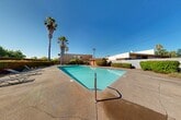 Located at 4730 El Camino Ave in Arden-Arcade, California, this single-level property features a generously sized outdoor swimming pool as its centerpiece. The pool area is surrounded by a wide concrete deck with poolside lounge chairs, offering ample space for relaxation and recreation. Towering palm trees and well-maintained hedges and shrubs frame the perimeter, providing a degree of privacy while enhancing the California outdoor aesthetic.

The adjacent building showcases a distinctive mid-century architectural style, characterized by a sawtooth roofline with angled wood beams and expansive floor-to-ceiling windows that allow abundant natural light to fill the interior. The warm-toned exterior combines wood accents with neutral stucco siding, creating a cohesive and inviting facade that overlooks the pool courtyard.

A small red outbuilding near the pool serves as an accessory structure, likely housing pool equipment or storage. Seating areas with tables and chairs are positioned along the building's exterior, ideal for outdoor gatherings. The landscaping throughout the space includes a variety of shrubs, ornamental plants, and mature trees, contributing to a vibrant and well-kept environment. The site benefits from the region's characteristic abundant sunshine, making this outdoor amenity a standout feature of the property.