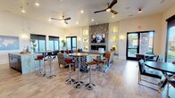 This single-floor space is designed as an inviting communal lounge or clubhouse, thoughtfully laid out to accommodate both relaxed gatherings and casual socializing. The open-plan interior is anchored by a striking floor-to-ceiling stone fireplace, which serves as the room's focal point and is flanked by illuminated built-in wall niches that add architectural depth and a warm, ambient glow. A wall-mounted flat-screen television above the fireplace enhances the entertainment-ready atmosphere.

The seating area is generously appointed with a mix of plush sofas, patterned accent chairs, and leather seating arranged around a glass-topped coffee table, creating a comfortable and stylish conversation space. Complementing this arrangement, high-top bar tables with chrome stools are positioned toward the front of the room, offering an alternative setting for more casual interaction.

Light wood-toned flooring runs throughout the space, contributing to an airy and contemporary aesthetic. Recessed lighting and ceiling fans with sleek wooden blades ensure comfort and even illumination across the entire area. Large windows and a glass door allow natural light to flood the interior, with views of a landscaped outdoor area visible beyond. The overall ambiance is modern, polished, and welcoming.