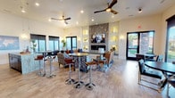 This single-floor space is designed as an inviting communal lounge or clubhouse, thoughtfully laid out to accommodate both relaxed gatherings and casual socializing. The open-plan interior is anchored by a striking floor-to-ceiling stone fireplace, which serves as the room's focal point and is flanked by illuminated built-in wall niches that add architectural depth and a warm, ambient glow. A wall-mounted flat-screen television above the fireplace enhances the entertainment-ready atmosphere.

The seating area is generously appointed with a mix of plush sofas, patterned accent chairs, and leather seating arranged around a glass-topped coffee table, creating a comfortable and stylish conversation space. Complementing this arrangement, high-top bar tables with chrome stools are positioned toward the front of the room, offering an alternative setting for more casual interaction.

Light wood-toned flooring runs throughout the space, contributing to an airy and contemporary aesthetic. Recessed lighting and ceiling fans with sleek wooden blades ensure comfort and even illumination across the entire area. Large windows and a glass door allow natural light to flood the interior, with views of a landscaped outdoor area visible beyond. The overall ambiance is modern, polished, and welcoming.