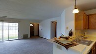 This single-floor space presents an open, functional layout with a seamless flow between the kitchen and living areas. The living room is finished with wall-to-wall carpeting in a neutral gray tone, contributing to a calm and understated ambiance. White walls throughout keep the space feeling bright and clean, while the standard ceiling height provides a comfortable sense of enclosure.

A peninsula-style counter separates the kitchen from the living area, offering a practical surface for dining or food preparation. Decorative vases and place settings on the counter add a modest touch of character. A pendant light above the counter provides focused task lighting in this zone.

The front entry is visible from the living area, featuring a dark wood door with an arched transom window that allows a small amount of natural light into the entryway. A floor vent near the entry wall indicates a forced-air heating system. Additional doorways lead to other areas of the unit, suggesting at least one additional room beyond the main living space. The overall design is straightforward and practical, suited for everyday living.