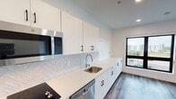 This single-floor unit showcases a contemporary design with clean lines and a neutral color palette throughout. The kitchen serves as a standout feature, equipped with stainless steel appliances including a built-in microwave, dishwasher, and refrigerator. White flat-front cabinetry with matte black hardware extends from floor to ceiling, providing ample storage, while a crisp white quartz countertop runs the full length of the galley-style layout. A herringbone-patterned tile backsplash adds visual texture and sophistication to the space. Wood-look plank flooring runs consistently throughout the unit, creating a cohesive and warm aesthetic.

The living area is open and airy, benefiting from a large picture window framed in black that invites natural light and offers views of the surrounding neighborhood. An additional low cabinet beneath the window provides built-in storage. A sliding barn door with a dark metal rail leads to a walk-in closet, combining practicality with a modern design element. Recessed lighting illuminates each room evenly. The bathroom maintains the unit's consistent design language, featuring white cabinetry, a single-basin vanity with a matching white countertop, a wall-mounted mirror, and dark wood-look flooring.