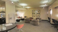 This single-floor lounge space is thoughtfully designed to provide a comfortable and inviting atmosphere for its occupants. The layout features a well-curated mix of contemporary furnishings, including curved upholstered club chairs in a warm taupe finish, a coordinating sofa with coral accent pillows, and a distinctive coral modern accent chair that adds a vibrant pop of color to the neutral palette. Soft, patterned carpet in muted grey tones grounds the space and adds texture underfoot. Overhead, recessed lighting and adjustable track lighting work in tandem to cast a warm, ambient glow throughout the room, complemented by a classic floor lamp near the seating area. The walls are painted in a soft, neutral beige tone, serving as a clean backdrop for a carefully arranged gallery of four framed art prints, displayed in a symmetrical two-by-two configuration. A low coffee table anchors the central seating arrangement, encouraging conversation and interaction. The overall ambiance is polished yet approachable, balancing modern design sensibilities with a sense of comfort, making this space well-suited as a professional lounge, waiting area, or communal gathering spot.