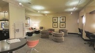 This single-floor lounge space is thoughtfully designed to provide a comfortable and inviting atmosphere for its occupants. The layout features a well-curated mix of contemporary furnishings, including curved upholstered club chairs in a warm taupe finish, a coordinating sofa with coral accent pillows, and a distinctive coral modern accent chair that adds a vibrant pop of color to the neutral palette. Soft, patterned carpet in muted grey tones grounds the space and adds texture underfoot. Overhead, recessed lighting and adjustable track lighting work in tandem to cast a warm, ambient glow throughout the room, complemented by a classic floor lamp near the seating area. The walls are painted in a soft, neutral beige tone, serving as a clean backdrop for a carefully arranged gallery of four framed art prints, displayed in a symmetrical two-by-two configuration. A low coffee table anchors the central seating arrangement, encouraging conversation and interaction. The overall ambiance is polished yet approachable, balancing modern design sensibilities with a sense of comfort, making this space well-suited as a professional lounge, waiting area, or communal gathering spot.