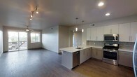 This single-floor space showcases a thoughtfully designed open-concept layout that seamlessly connects the kitchen and living area. The kitchen is a standout feature, equipped with stainless steel appliances including a range, microwave, and dishwasher. White shaker-style cabinetry extends to the ceiling, complemented by a subtle tile backsplash in a mosaic pattern that adds texture without overwhelming the clean aesthetic. A peninsula with a white countertop and undermount sink serves as both a functional workspace and a natural divider between the kitchen and the adjoining living area, with pendant lighting hanging above to define the space. Recessed lighting and a track light fixture provide ample illumination throughout. The flooring transitions between wood-tone planks, lending warmth and continuity across the open floor plan. The living area benefits from a window that allows natural light to fill the space, creating a bright and welcoming atmosphere. Neutral wall tones and a consistent design palette give the space a cohesive, modern feel throughout.