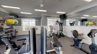 This fitness center offers a thorough and well-organized workout environment designed to accommodate a wide range of exercise routines. The space is finished with a cool blue-grey color palette, white wainscoting, and crisp crown molding, lending it a clean and polished aesthetic. Durable rubber flooring covers the entire area, providing both safety and functionality underfoot.

Natural light streams in through multiple large, multi-pane windows that frame outdoor greenery, creating an open and refreshing atmosphere. Ceiling fans and recessed panel lighting ensure the space remains comfortable and well-lit throughout. Wall-mounted flat-screen televisions are positioned at multiple points around the room for convenience during workouts.

Large mirrors line a portion of the walls, visually expanding the space and allowing users to monitor their form. The equipment selection is extensive, including treadmills, recumbent and upright stationary bikes, an elliptical, and a rowing machine for cardiovascular training. Strength training is well-served by multiple selectorized cable and weight machines, a power tower, and a dedicated free weights area. The free weights section features a fully stocked rack of dumbbells, kettlebells, medicine balls, stability balls, battle ropes, and a heavy punching bag, supporting functional and high-intensity training as well.