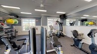 This fitness center offers a thorough and well-organized workout environment designed to accommodate a wide range of exercise routines. The space is finished with a cool blue-grey color palette, white wainscoting, and crisp crown molding, lending it a clean and polished aesthetic. Durable rubber flooring covers the entire area, providing both safety and functionality underfoot.

Natural light streams in through multiple large, multi-pane windows that frame outdoor greenery, creating an open and refreshing atmosphere. Ceiling fans and recessed panel lighting ensure the space remains comfortable and well-lit throughout. Wall-mounted flat-screen televisions are positioned at multiple points around the room for convenience during workouts.

Large mirrors line a portion of the walls, visually expanding the space and allowing users to monitor their form. The equipment selection is extensive, including treadmills, recumbent and upright stationary bikes, an elliptical, and a rowing machine for cardiovascular training. Strength training is well-served by multiple selectorized cable and weight machines, a power tower, and a dedicated free weights area. The free weights section features a fully stocked rack of dumbbells, kettlebells, medicine balls, stability balls, battle ropes, and a heavy punching bag, supporting functional and high-intensity training as well.