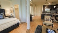 This single-floor residence at Broadstone Lincoln offers an open-concept layout that connects the living, dining, and sleeping areas in a cohesive and functional flow. Warm-toned hardwood floors extend throughout the space, contributing to a bright and welcoming ambiance that ties each zone together visually.

The sleeping area is positioned to one side, providing a sense of separation from the main living space while remaining part of the open floor plan. A walk-in closet is accessible directly from the bedroom area, offering generous storage with visible shelving and hanging space. A structural column adds an architectural element that subtly defines the transition between the bedroom and the central living and dining zones.

The dining area features a compact, round table with seating, positioned beneath track lighting that adds focused illumination. The kitchen is visible toward the rear of the space, highlighted by dark cabinetry that contrasts with the light-toned walls and flooring. The main living area accommodates seating and is accented by a patterned area rug.

Overall, the residence maintains a clean, contemporary aesthetic with soft neutral tones, efficient use of space, and a layout that promotes easy movement throughout.