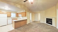 This single-story space showcases an open-concept layout that seamlessly connects the kitchen and living area, creating a cohesive and functional environment. The kitchen is equipped with white appliances, including an electric range and built-in microwave, complemented by warm honey-toned wooden cabinetry that provides ample storage. A peninsula with a light countertop extends into the living space, offering additional prep area and casual seating potential.

A defining architectural feature is the vaulted ceiling, which rises above both the kitchen and living area, enhancing the sense of openness and volume. A ceiling fan is centrally positioned to maintain airflow throughout. The living area is finished with plush, medium-toned carpeting that adds warmth underfoot.

Neutral, warm beige walls create a cohesive backdrop throughout the space. A window near the kitchen sink allows natural light to filter in. Toward the back of the space, multiple doorways lead to additional rooms, including what appears to be a closeted storage area, offering practical organization options. The overall ambiance is bright, functional, and inviting.