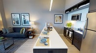 This single-floor residence showcases a thoughtfully designed open-concept layout that unifies the kitchen, dining, and living spaces into one cohesive environment. The kitchen is outfitted with stainless steel appliances, including a range and built-in microwave, complemented by dark cabinetry with open upper shelving for decorative storage. A long, white-topped island serves as the centerpiece of the space, doubling as both a food preparation area and a casual dining surface, with place settings arranged along its length to illustrate its versatility.

Light wood-tone flooring runs throughout, lending warmth to the otherwise cool, neutral gray walls. Recessed and linear LED lighting overhead provides consistent, modern illumination across the open floor plan. The adjoining living area features a dark upholstered sofa accented with colorful throw pillows, a warm-toned side table with a lamp, and framed artwork on the walls, contributing to an inviting atmosphere. The overall aesthetic balances clean, contemporary design with comfortable, livable details.