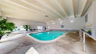 This indoor pool facility in Caledonia, Wisconsin offers a well-appointed aquatic environment designed for both recreation and relaxation. The centerpiece of the space is a generously sized swimming pool with vivid turquoise water, accessible via stainless steel entry rails and steps. Adjacent to the pool is a whirlpool spa, clearly marked with posted capacity and safety guidelines for guest use.

The architecture features a striking exposed beam ceiling with parallel structural ribs painted in a clean white finish, lending the space a sense of height and openness. The concrete pool deck provides ample circulation space around the water, while large windows and sliding glass doors along one wall flood the interior with natural light and offer views of the surrounding outdoor landscape.

The lounge area is furnished with a row of contemporary slatted chairs arranged along the windowed wall, complemented by an assortment of large potted tropical plants that add warmth and a resort-like ambiance to the interior. Wall-mounted sconces provide additional ambient lighting throughout the facility.

White tile walls and baseboard heating units line the perimeter, contributing to the facility's practical, well-maintained character. Safety signage, including pool and spa rules, is clearly posted. A staircase provides access to adjacent areas of the building, making this pool facility a functional and inviting recreational amenity.