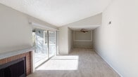 This single-floor residence offers a thoughtfully designed layout that balances comfort and functionality throughout. The living area is a standout feature, showcasing vaulted ceilings that lend an airy, open feel to the space. Large sliding glass doors flood the room with natural light and provide direct access to a private balcony with outdoor views. A ceiling fan adds both style and practicality to the adjoining dining or sitting area, which is defined by a subtle architectural step in the ceiling. A second sliding glass door leads to another outdoor space and frames a classic brick fireplace flanked by built-in shelving, creating a warm and inviting focal point. Soft carpeting flows throughout the main living spaces and bedrooms, adding warmth underfoot. The kitchen features white cabinetry, speckled laminate countertops, a double stainless steel sink, and black appliances including a dishwasher, all illuminated by a window overlooking the outdoors. Two well-sized bedrooms offer plush carpet, neutral tones, and windows with blinds for natural light and privacy. The bathrooms are cleanly finished with tile tub-surround combinations, vanities with speckled countertops, and framed mirrors. Textured ceilings are present throughout, adding a classic residential character to the entire home.